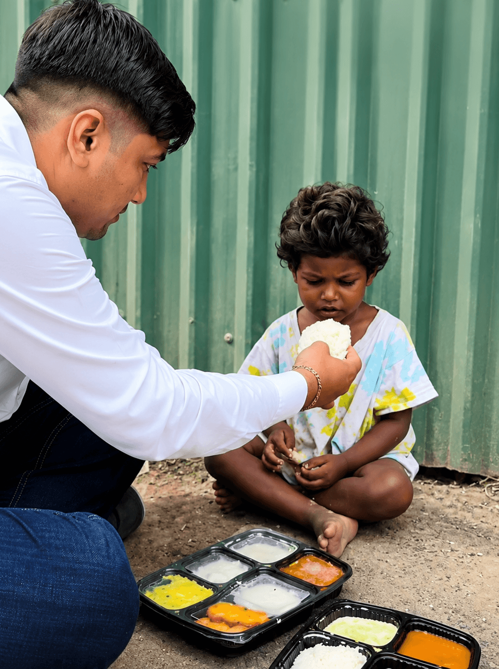 A volunteer kneels on the ground to offer rice from a packed meal tray to a child sitting in front of him.