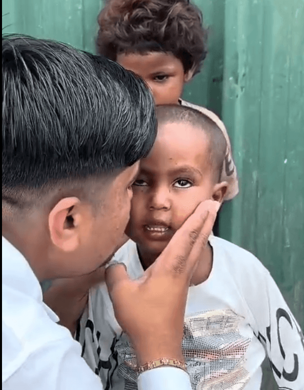 A volunteer holds a young child's cheek in one hand while a second child looks on from behind.