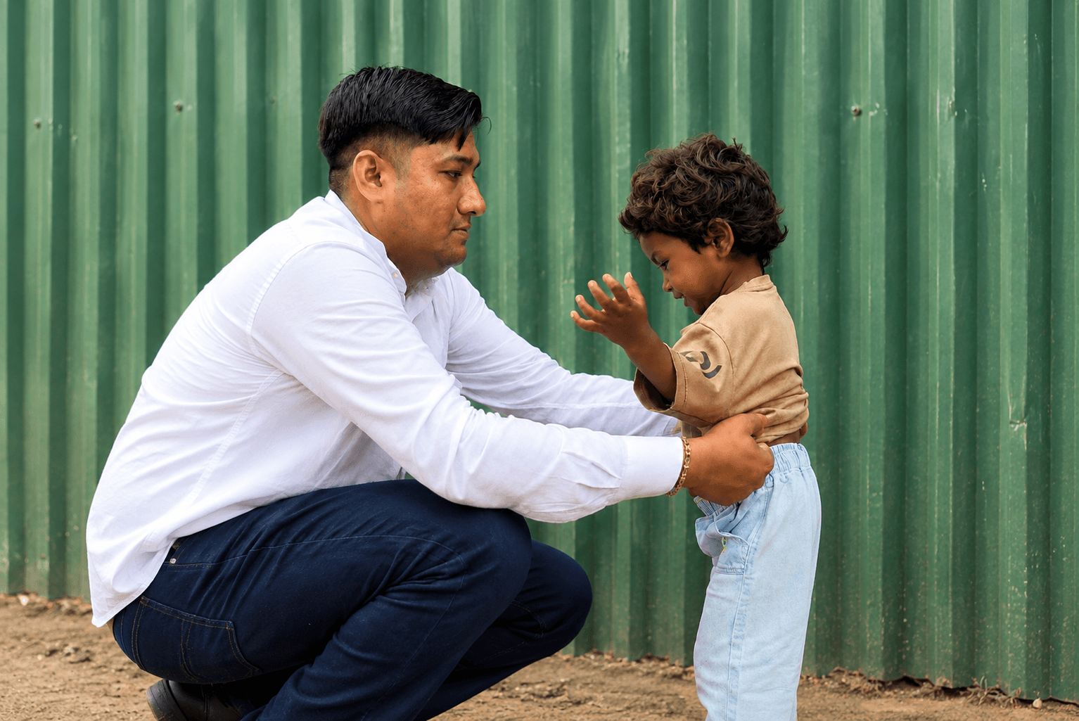 A volunteer crouches to steady a young child by the waist, the two of them face to face against a corrugated metal wall.