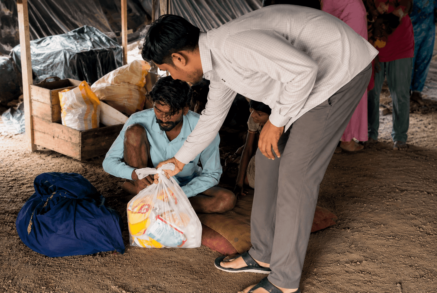 A volunteer leans down to hand a carrier bag of packaged provisions to a man seated on the ground under a tarpaulin shelter, with sacks and crates stacked behind them.